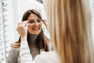 Fototapeta premium Beautiful and fashionable woman choosing eyeglasses frame in modern optical store. Female seller specialist helps her to make right decision.