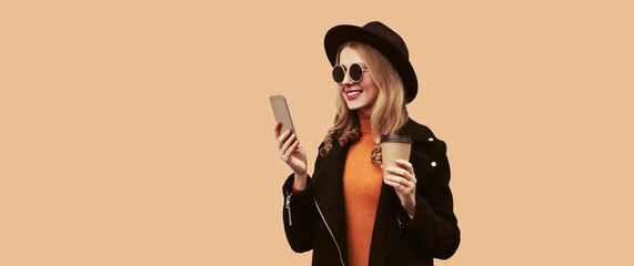 Portrait of stylish smiling woman looking at smartphone holding coffee cup wearing black coat, round hat isolated on brown background