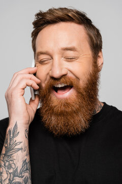 excited bearded man with closed eyes talking on mobile phone and laughing isolated on grey.