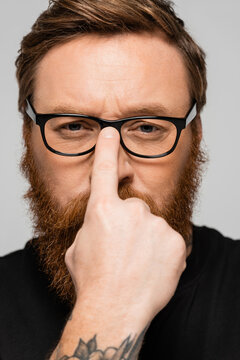 portrait of strict bearded man adjusting eyeglasses and looking at camera isolated on grey.