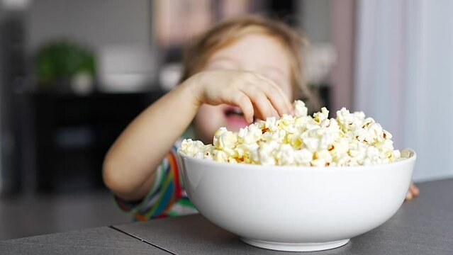 Little girl is eating popcorn in home kitchen. Focus on hand taking popcorn