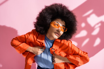 curly african american woman in stylish orange sunglasses and trendy outfit posing on pink.