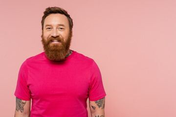 positive bearded and tattooed man in magenta t-shirt smiling at camera isolated on pink.