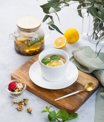Herbal tea with lemon and mint in a white cup on a wooden plate on a light background with eucalyptus branches and a teapot.