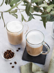Two cappuccino or latte with milk foam in glasses on a light background with coffee beans and eucalyptus branch.
