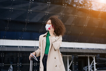 Young african woman girl in wearing face mask ffp2 walking outdoors carrying a suitcase and going...