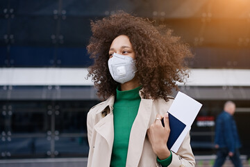 Young african woman girl in wearing face mask ffp2 walking outdoors carrying a suitcase and going...