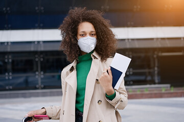 Young african woman girl in wearing face mask ffp2 walking outdoors carrying a suitcase and going...