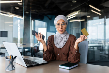 Worried and upset young muslim businesswoman in hijab holding phone and credit card. Sitting in the office at the desk, looking at the camera.