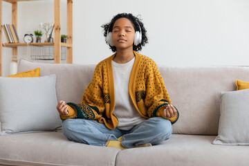 Obraz premium Yoga mindfulness meditation. Young healthy african girl practicing yoga at home. Woman sitting in lotus pose on couch meditating smiling relaxing indoor. Girl doing breathing practice. Yoga at home