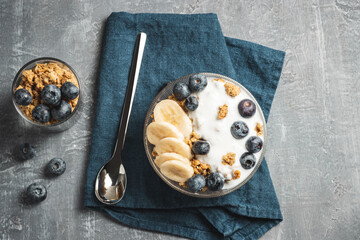 Granola cereal oatmeal with white yogurt, blueberries and banana fruits in a bowl on a blue napkin, grey background, top view