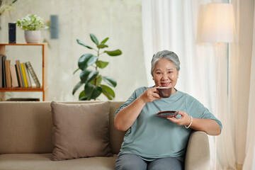 Portrait of happy aged woman drining cup of coffee and watching movie on tv