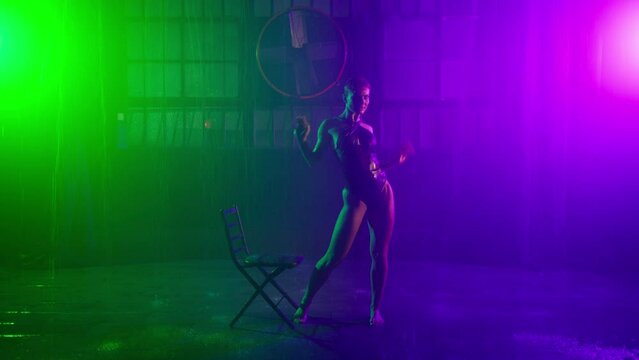 Young hot woman with short pink hair performs sensual strip dance using chair in room with falling rain and having wet body from water droplets falling from above, slow motion colorful footage on RED