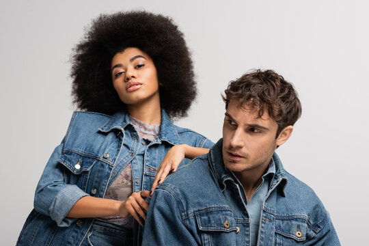 brunette african american woman looking at camera and leaning on man in denim outfit isolated on grey.