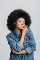 curly african american woman in denim jacket looking at camera while posing isolated on grey.