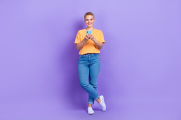 Fototapeta na wymiar Full size photo of cheerful pretty lovely girl dressed sneakers denim pants holding smartphone isolated on purple color background