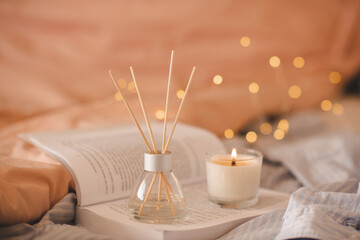 Liquid home fragrance in glass bottle and bamboo sticks with scented candles on open paper book in bed over glow lights closeup. Cozy atmosphere. Good morning.
