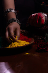 Young Indian girl woman in ethnic attire saree and bangles in hand playing with organic dry color powder gulal abeer in a clay plate celebrating Holi festival of colors with smoke in the background