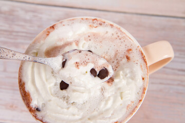 Cup of hot chocolate with whipped cream sprinkled with chocolate chips, being stirred with a spoon, on a wooden background
