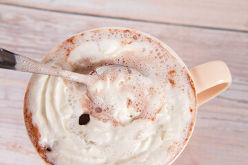 Cup of hot chocolate with whipped cream sprinkled with chocolate chips, being stirred with a spoon,...