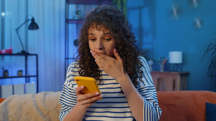 Happy woman with curly hairstyle use mobile smartphone typing browsing say Wow yes found out great big win good news celebrate lottery jackpot doing winner gesture. Girl at home in room sits on sofa