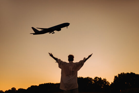 Silhouette Of A Girl With Outstretched Arms Under An Airplane Taking Off At Sunset In Summer. The Girl Under The Plane Dreams Of Rest. Watching The Plane Take Off.