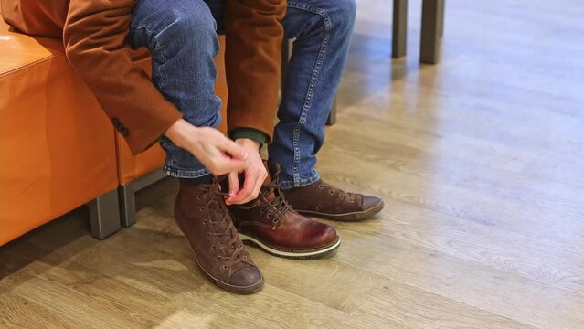 A young handsome caucasian brunette in a brown coat takes off an old shoe to try on a new one, sitting on a leather pouffe in a shoe store, close-up side view. The concept of offline shopping, men's l