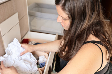 Young pregnant mother lovingly arranges her baby clothes in the drawer chest