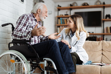 Young nurse talking with sad senior man in a wheelchair during home visit
