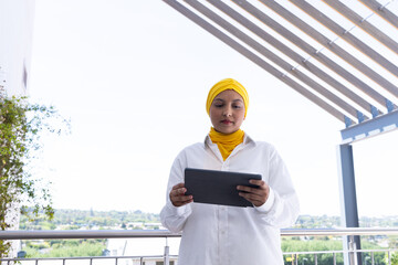 Happy biracial businesswoman with hijab using tablet outside office