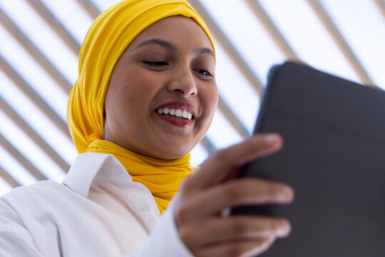 Happy biracial businesswoman with hijab using tablet outside office