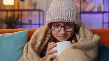 Sick toddler girl wear hat wrapped in plaid sit alone shivering from cold on sofa drinking hot tea...