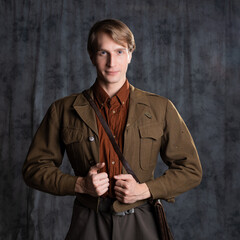 Fototapeta na wymiar A young guy in military-style clothes, a brown flight jacket and breeches with suspenders. Posing in the studio on a gray background, close-up