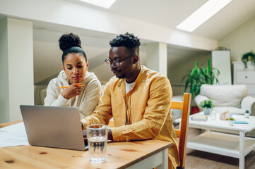 Diverse couple using laptop while sitting at the table at home