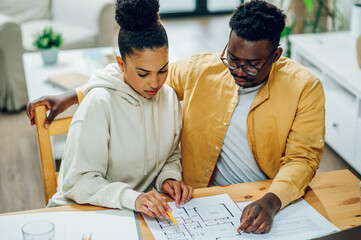 Diverse couple using laptop and looking into the blueprints of their new home