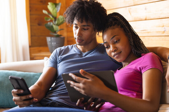 Happy african american couple spending time in log cabin using tablet and smartphone
