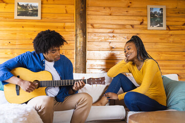 Happy african american couple spending time in log cabin playing guitar