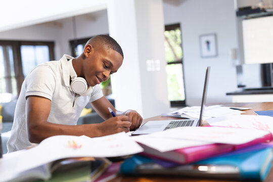 Image of happy african american teenage boy doing homework with laptop at home