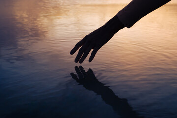 Female hand touching the calm water ocean lake surface reflecting a beautiful summer sunset