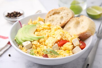 Bowl with delicious scrambled eggs, tofu, avocado and slices of baguette on white table, closeup