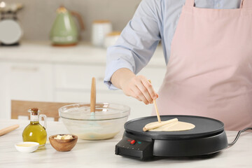 Woman cooking delicious crepe on electric pancake maker at white marble table in kitchen, closeup