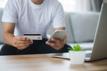 Man sitting on sofa hand holding smart phone paying with credit card on smart phone at home. He used to pay by credit card on a regular basis as it was convenient and he didn't have to carry cash.