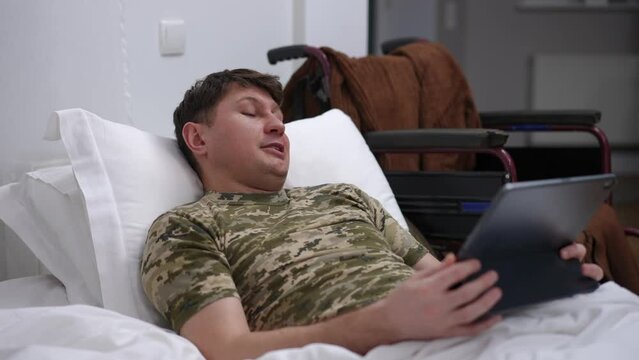 Medium shot Caucasian man lying in bed talking at video chat in digital tablet application with blurred wheelchair at background. Portrait of positive courageous soldier messaging on rehabilitation