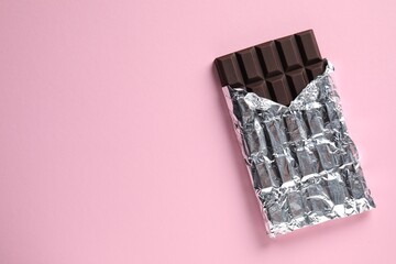 Delicious dark chocolate bar wrapped in foil on pink background, top view. Space for text
