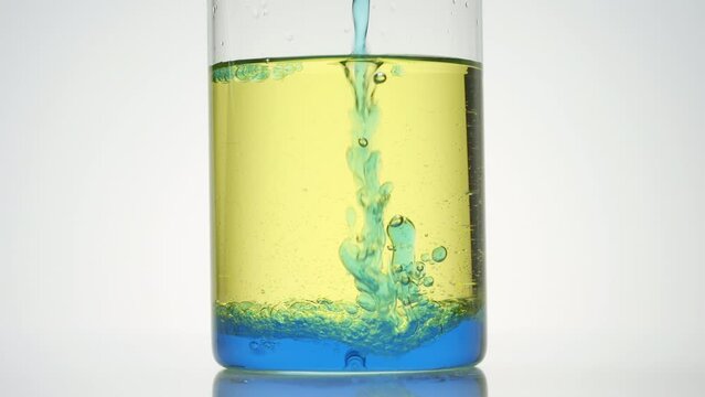 Side view macro shot of blue oil is being poured into beaker with blue and yellow oils on grey background | Abstract body care cosmetics ingredients formulating concept