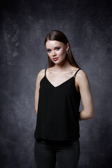 Portrait of female in black tank top with straps and jeans. Woman with earrings in the ear on gray background.