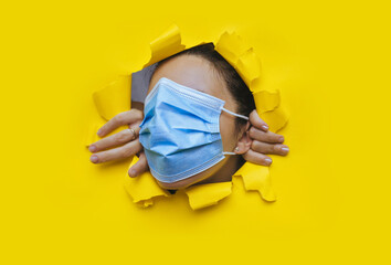A woman wearing a protective medical mask that completely covers her face peeps through a torn hole...