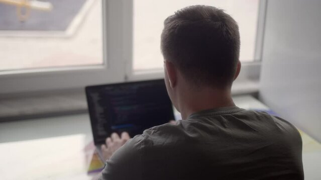 Rear view of man who works on laptop, programmer. IT specialist at home office work in front of bright light window. professional web designer programs back end site.