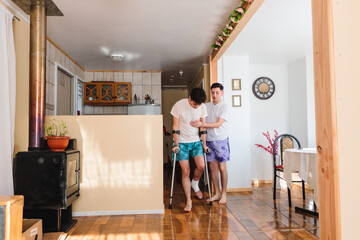 Homosexual man in pajama supporting his injured gay partner who is using crutches indoors. LGBT...