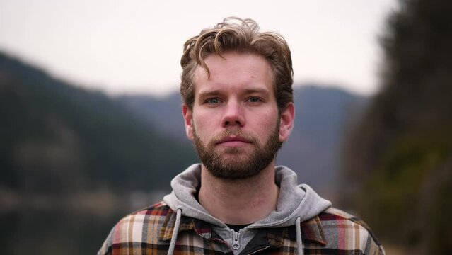 A serious looking man with a little winker. His blond wavy hair fits perfectly. In the backround is a cloudy hill full of trees and a small part of a river is visible behind the man.